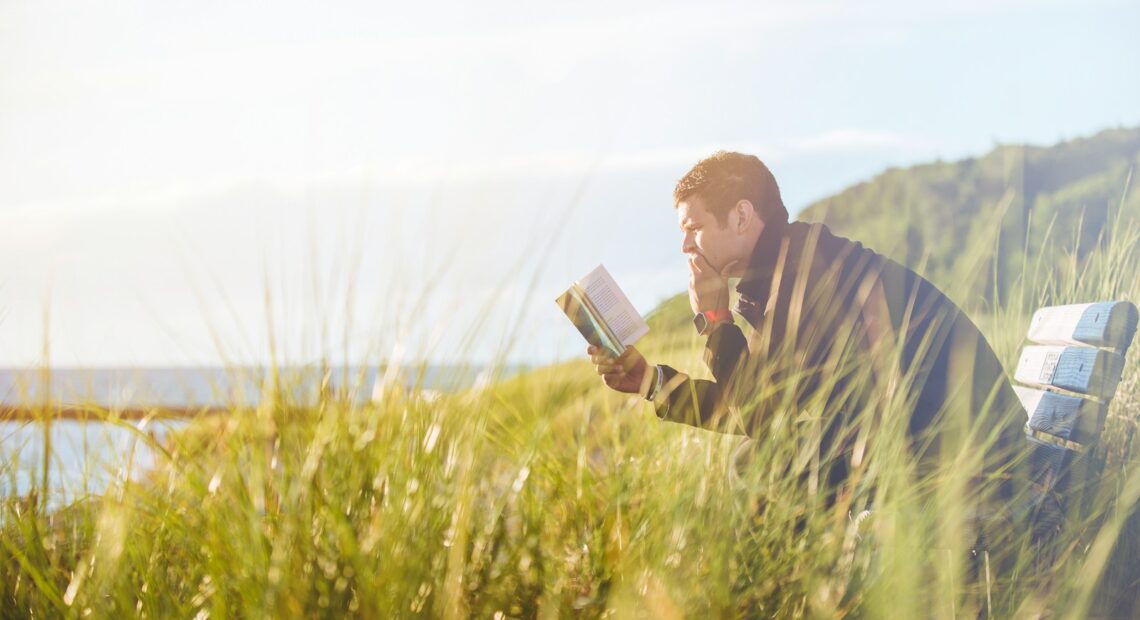 man reading book on beach near lake during daytime