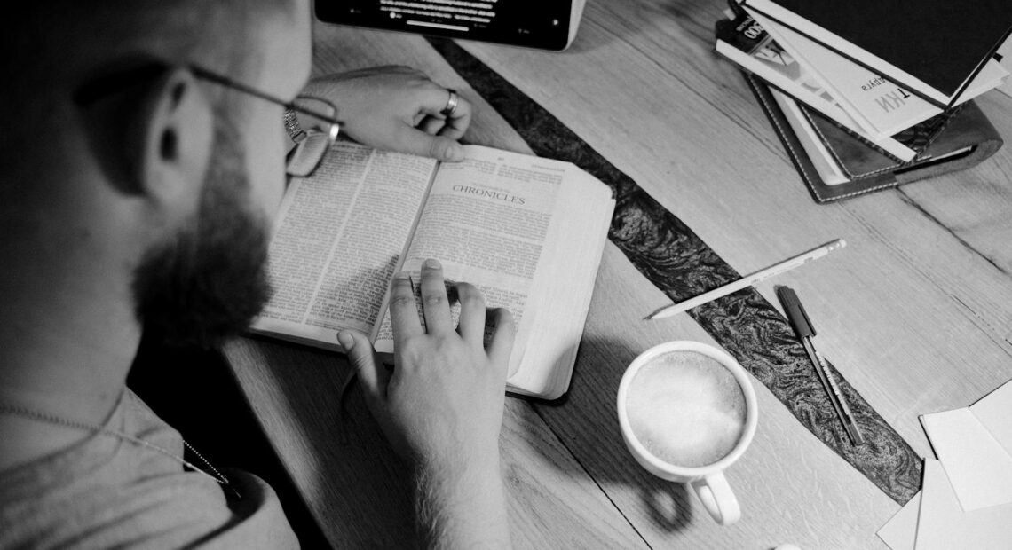 A man reading a bible with a coffee cup, books, and stationery on a wooden table.