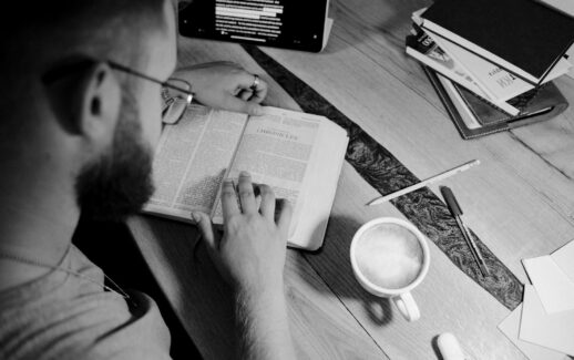 A man reading a bible with a coffee cup, books, and stationery on a wooden table.
